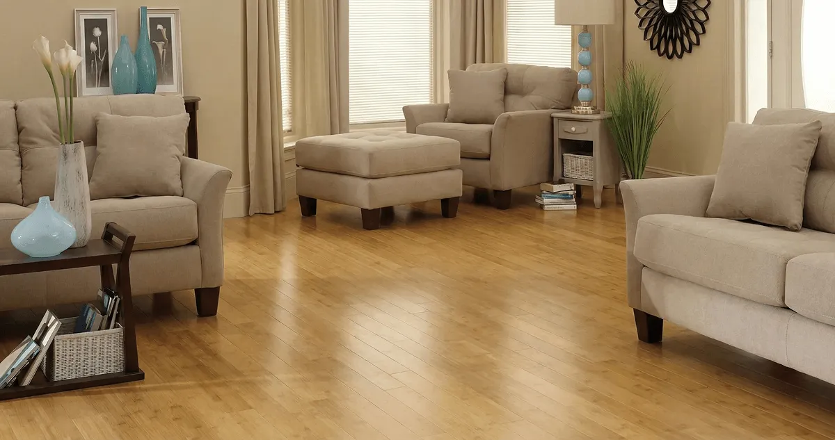 A living room with professionally installed bamboo flooring, beige sofas, a matching ottoman, a side table with a blue lamp, vases, framed pictures, and large windows with blinds and curtains in Shreveport, LA