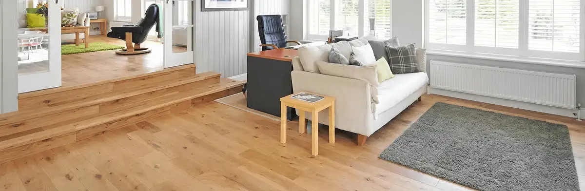 A modern living room with professionally installed laminate flooring, a beige sofa, a small side table, a gray rug, and large windows with white shutters in Shreveport, LA